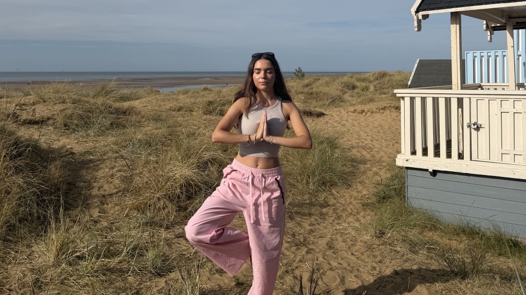 Young woman at beach doing yoga three pose and prayer. Wearing pink linen trousers.