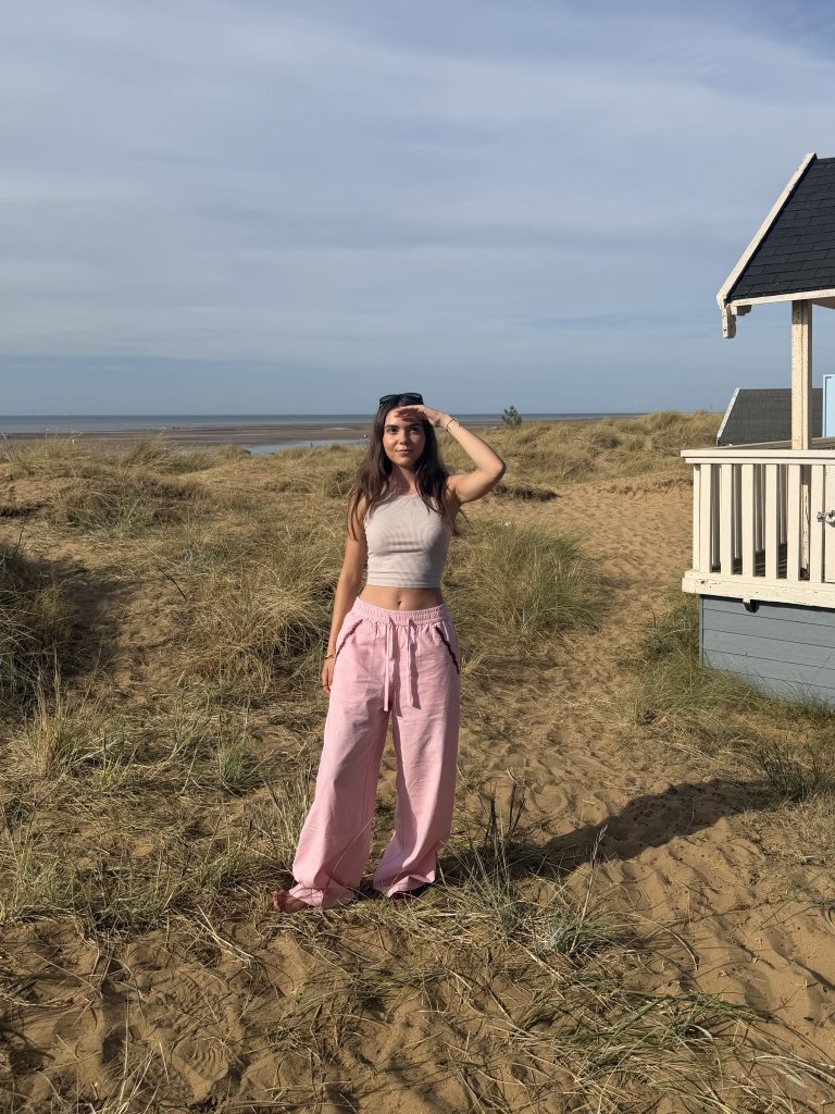 Young woman with long dark hair standing at the beach leading up to a teal beach hut, wearing a beige crop top and wide-leg pink trousers, with sandy dunes and coastal grass in the background