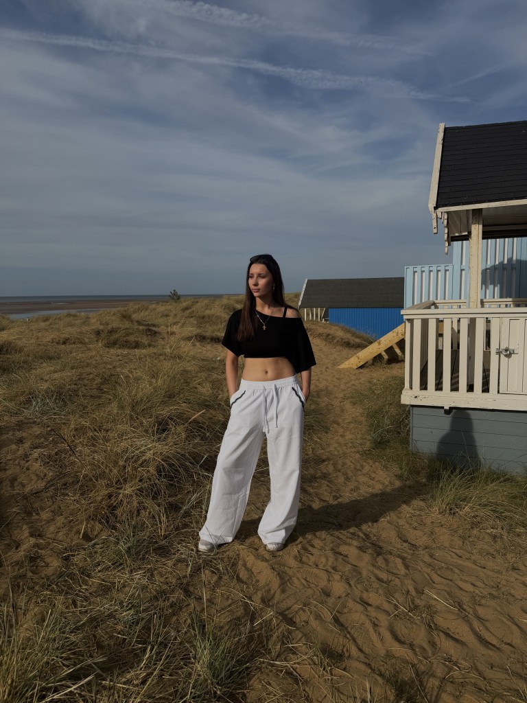 A young woman with long dark hair stands on sandy dunes among dry grass, wearing a black off-shoulder crop top and wide-leg white trousers, with colourful beach huts and a vast coastal horizon visible behind her.
