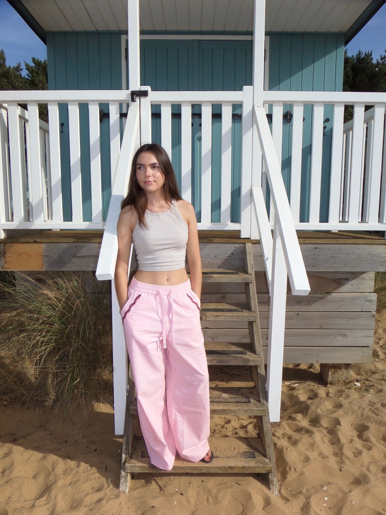 Young woman with long dark hair standing at the base of wooden steps leading up to a teal beach hut, wearing a grey crop top and wide-leg pink trousers, with sandy dunes and coastal grass in the background