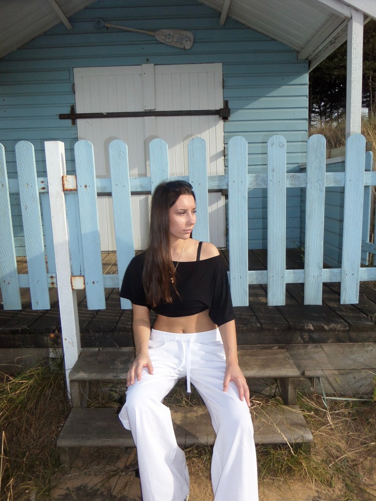 A young woman with long dark hair sits on the wooden steps of a light blue beach hut, wearing a black off-shoulder crop top and white wide-leg trousers, gazing to the side with sunglasses resting on her head.